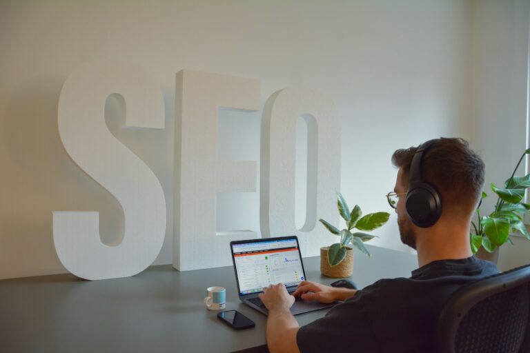 A man in headphones working on a laptop at a desk in front of large white SEO letters