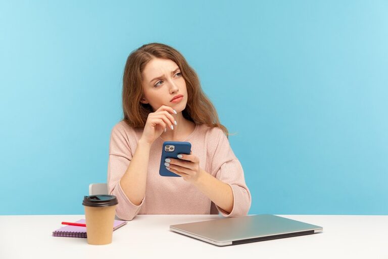 A thoughtful woman holding her phone at a desk with a coffee and a laptop