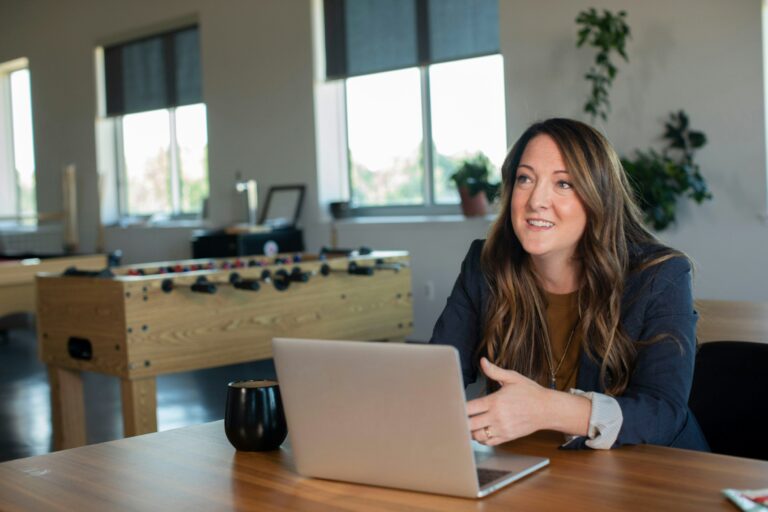 A smiling woman talking across a table with her laptop open in a bright office