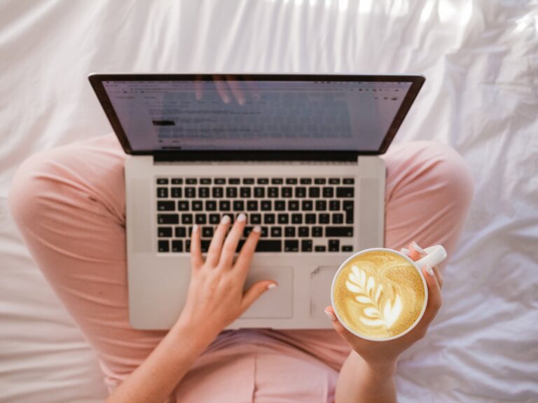 Someone sitting on a bed with a laptop on their lap and a latte in hand